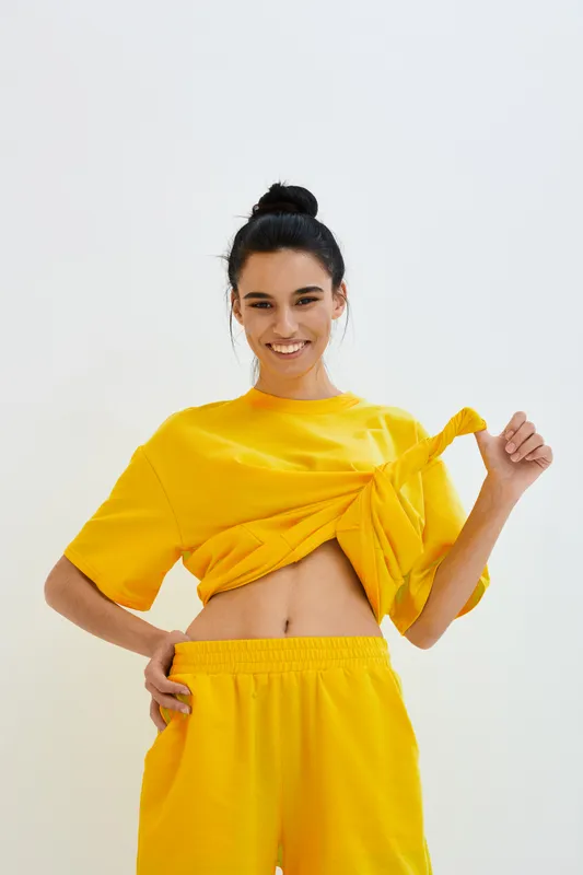 Young smiling woman in yellow casual outfit lifting top, showing toned stomach against plain light background, radiating energy and freshness in bright sporty style.