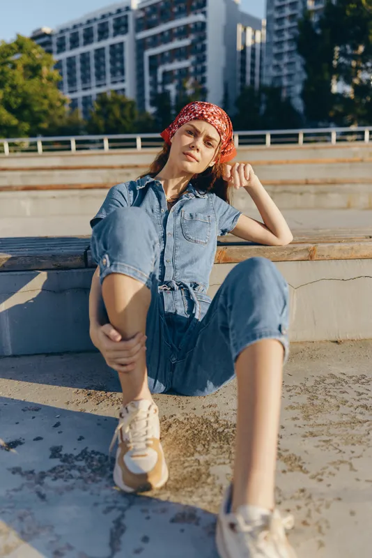 relaxed woman sitting outdoors in denim clothes and red bandana with casual sneakers on urban concrete steps under sunlight