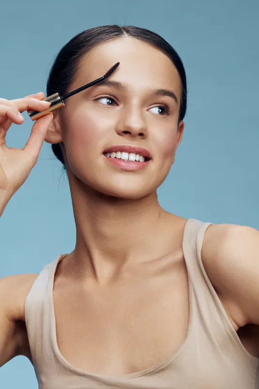 Smiling young model applying eyebrow gel with a soft expression against a light blue background, showcasing natural beauty and confidence