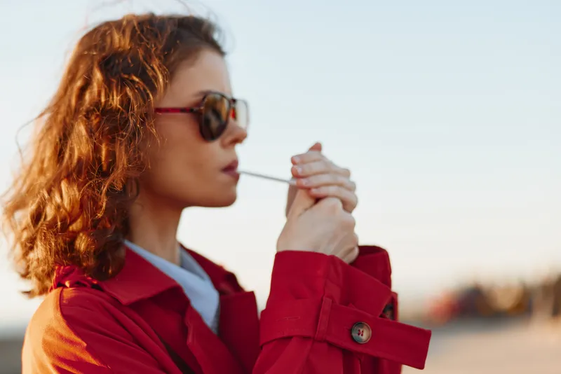 Young woman with red hair in a red trench coat lights a cigarette outdoors, wearing sunglasses, capturing a calm cinematic moment with natural light, gentle breeze, and confident posture against