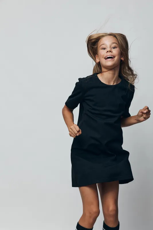 Girl, child, smiling and jumping in black dress on plain background. Happy kid with long hair, playful and energetic for childhood, fashion and lifestyle indoors.