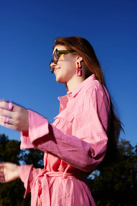 Cheerful young woman in a pink shirt dress enjoying a sunny day outdoors with stylish sunglasses and accessories, embodying summer fashion vibes