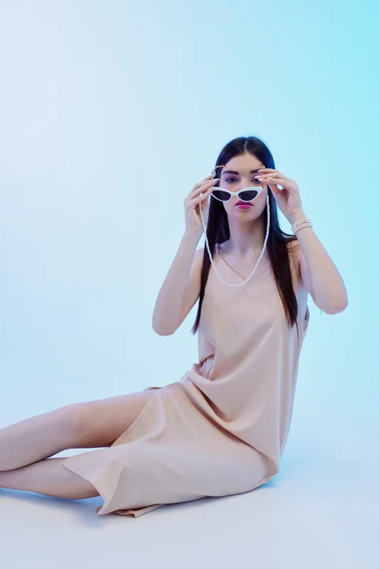 Young woman in a beige dress adjusts her sunglasses while sitting against a light blue background. Concept of beauty, fashion, and modern style with elegant accessories.