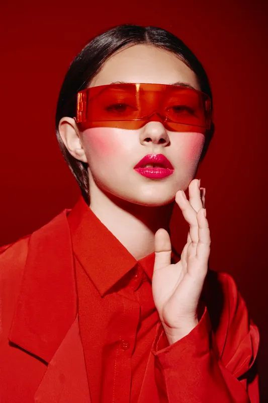 Fashion model with red sunglasses and vibrant red outfit poses against red background. Glamorous makeup complements creative beauty concept. Stylish portrait shot with high contrast lighting.