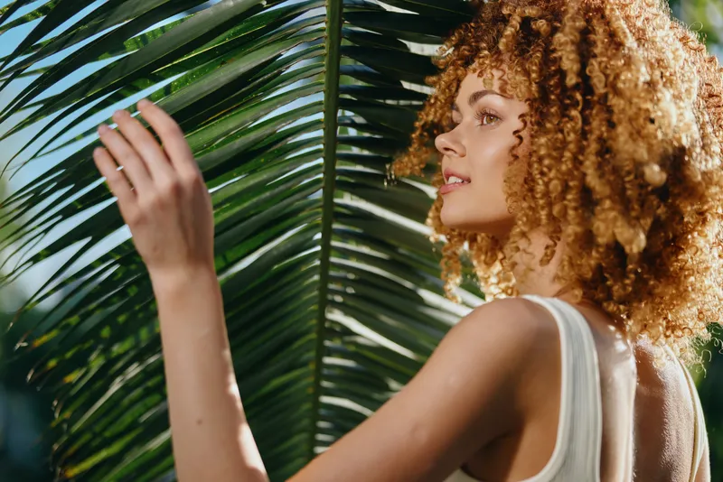 Curly woman touching large tropical palm leaf outdoors in sunlight, wearing white tank top, natural beauty with healthy skin and confident expression in summer setting.