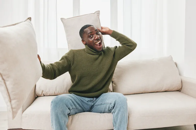Happy African man playfully relaxing on a couch with pillows, wearing a cozy green sweater and jeans, showcasing a joyful expression in a bright indoor setting