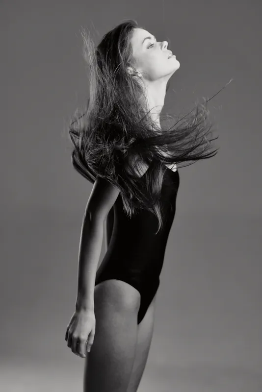 Black and white portrait of a young woman with flowing hair in a sleeveless bodysuit, expressing elegance and motion in studio lighting.