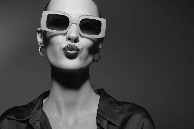 Fashionable white woman with light skin wearing large sunglasses and hoop earrings makes a kiss face on grey background.