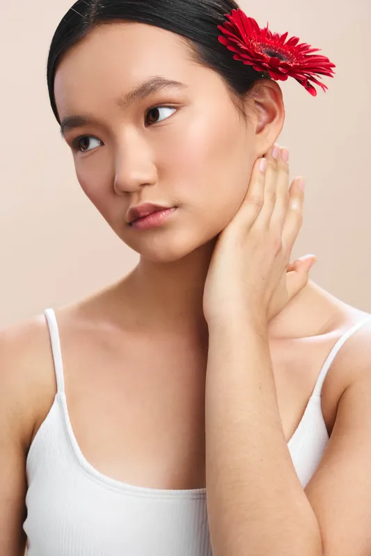 Asian woman portrait with a red flower in her hair, wearing a white sleeveless top. Soft lighting and a calm, graceful pose convey serenity, elegance, and refined beauty in a studio setting.