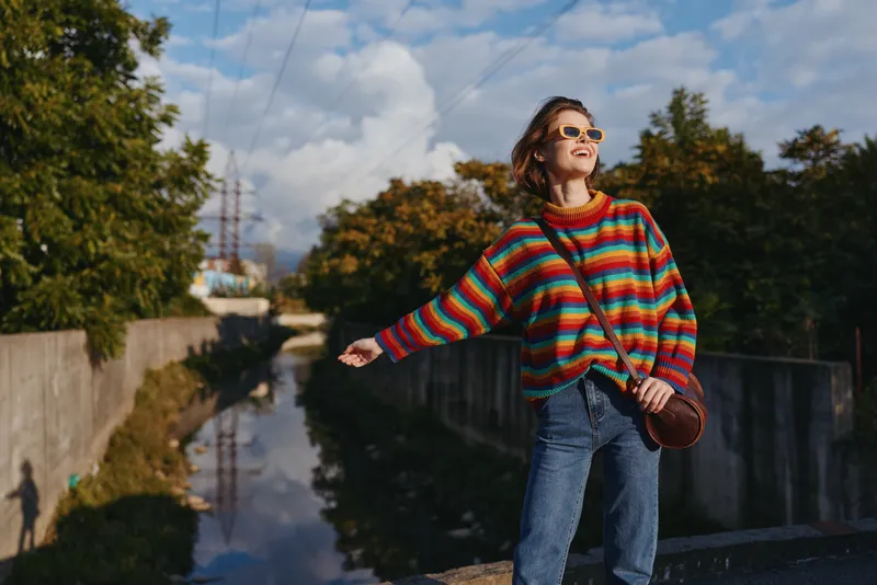 Woman in a colorful striped sweater and sunglasses, smiling and leaning on a bridge over a canal in autumn; casual jeans, leather crossbody bag, relaxed urban travel vibe.