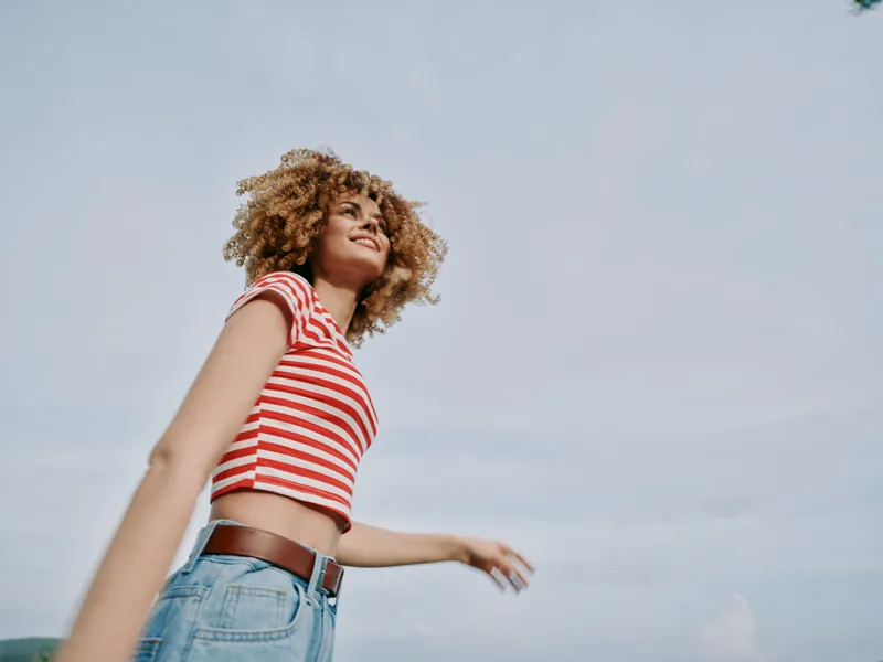 Young woman with curly hair wears a red and white striped crop top and denim jeans, walking outdoors with a carefree smile under a bright sky, expressing freedom.