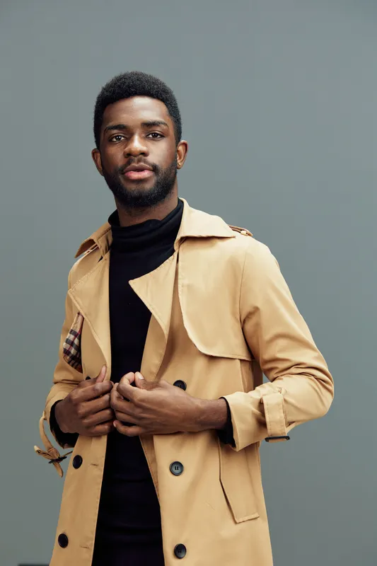 Stylish young Black man wearing a beige trench coat over a black turtleneck, expressing confidence against a muted gray background