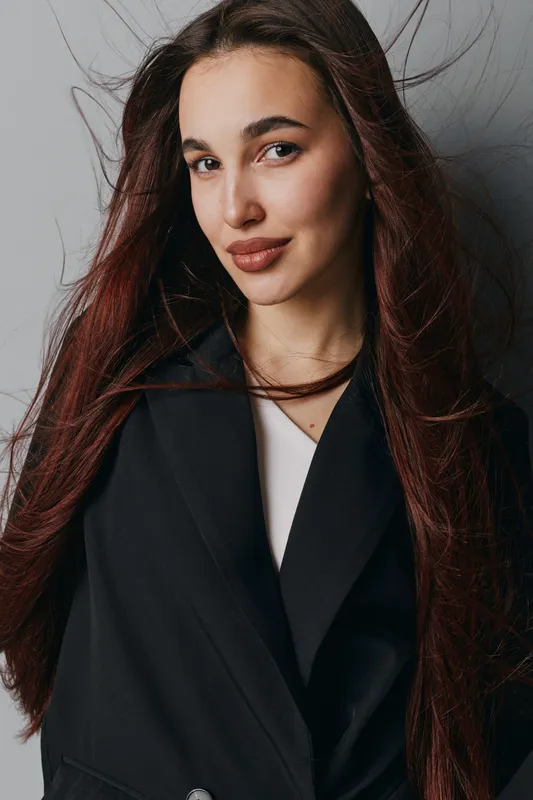Young woman with long brown hair wearing black coat posing with slight smile near white wall. Natural makeup, confident look, indoor portrait with casual hairstyle and soft lighting.