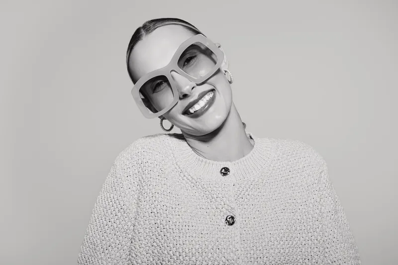 Smiling white woman with pale skin and slicked back hair wears large sunglasses and textured sweater. Studio portrait with fashionable and confident female model expression.