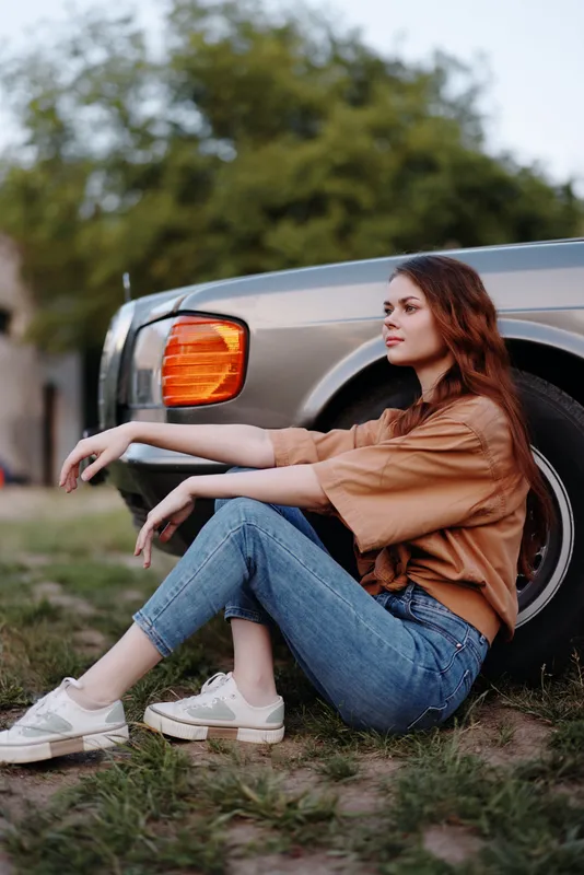 Young woman sitting by a classic car, dressed in casual attire, exuding a relaxed vibe with lush greenery in the background, perfect for lifestyle and vintage themes.