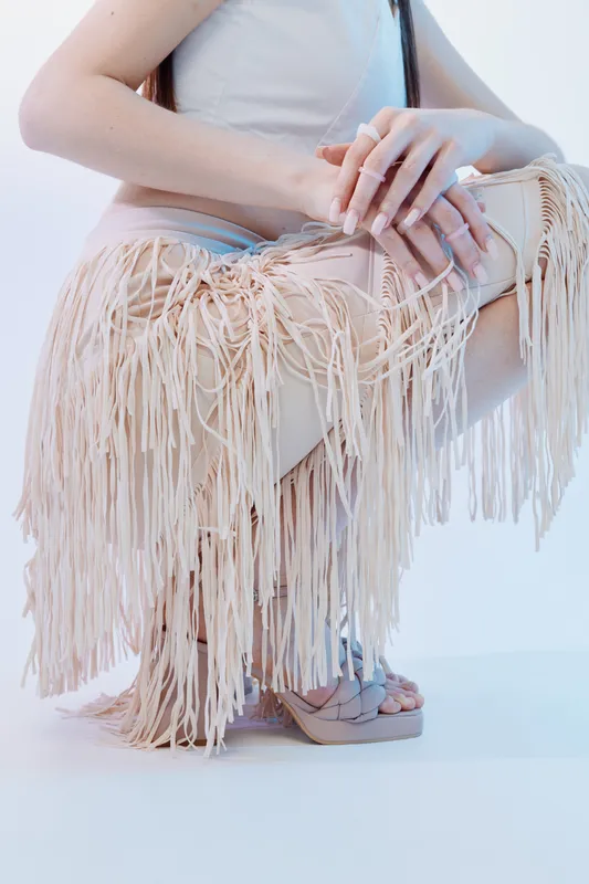 Woman beauty fashion concept featuring a close-up of hands holding a fringed beige handbag, wearing light clothing and sandals on a white background.