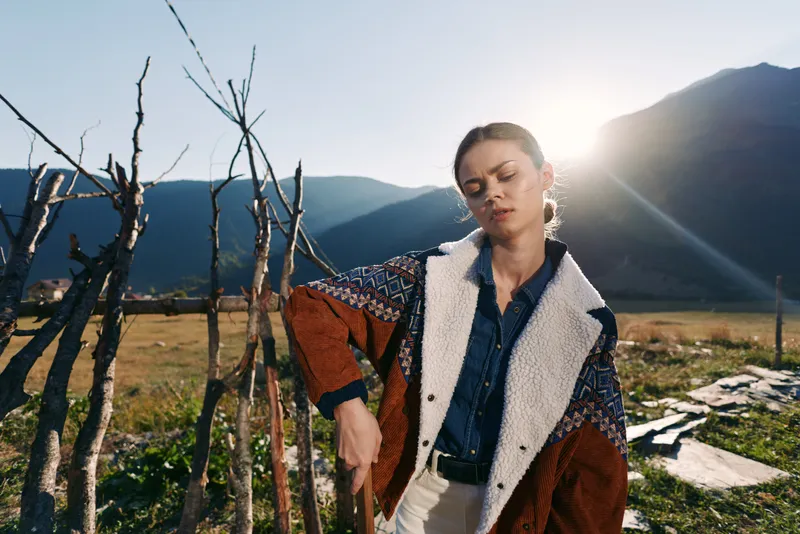 Woman portrait in mountains countryside at golden hour with sun backlight, wearing denim shirt and patterned jacket with shearling by a rustic fence in a serene outdoor field landscape.