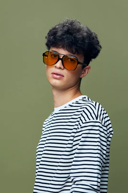 Young man with curly hair wearing large orange sunglasses and a striped shirt standing against a solid green background with a confident expression.