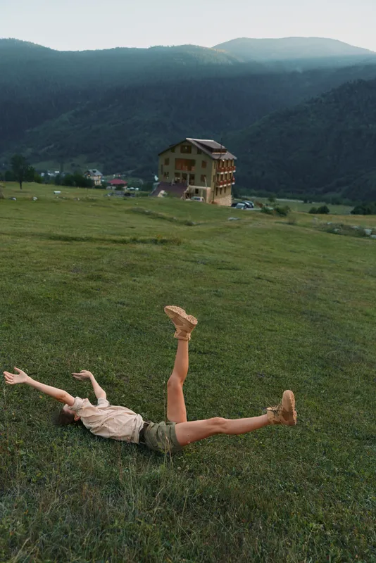 A woman in casual clothes lies on green grass with arms and legs raised in a mountainous rural landscape near a wooden house. Concept of lifestyle and freedom.