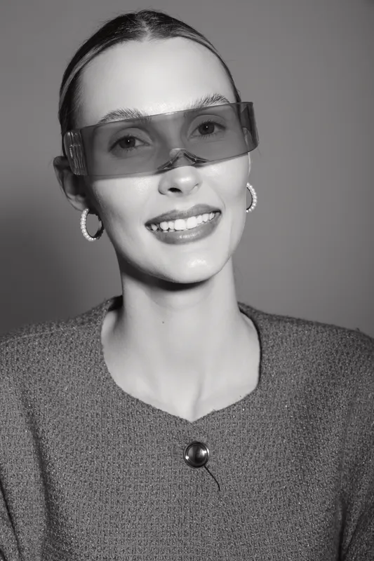 Young caucasian woman with fair skin wearing futuristic sunglasses and hoop earrings smiles confidently in studio portrait. Concept of fashion and style.