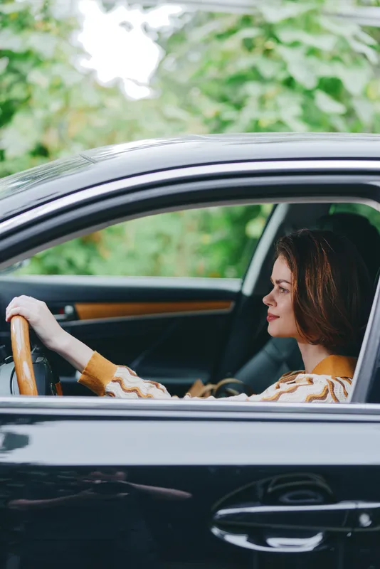 A woman driver sits behind the wheel of a sleek car, hands on the steering wheel, focused expression amid daytime greenery.