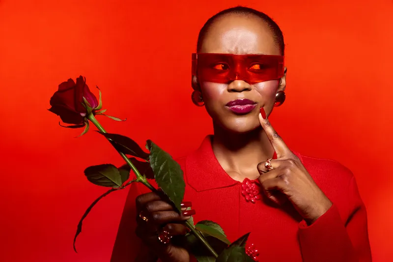 Portrait of an elegant dark skinned African woman wearing red sunglasses and a red sweater holding a red rose on a red background. Concept for fashion, style, and beauty expression.