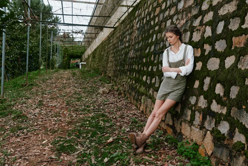 A thoughtful woman in casual attire stands against a rustic stone wall, surrounded by nature, evoking a serene outdoor vibe, ideal for lifestyle themes