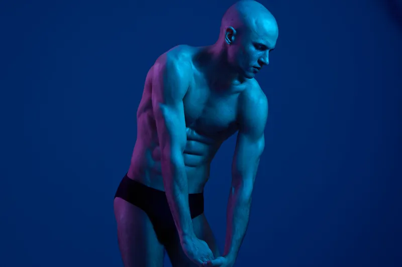 Stylish man posing in a dramatic blue light, showcasing his toned physique and athletic features. The image captures strength and confidence in a striking way.