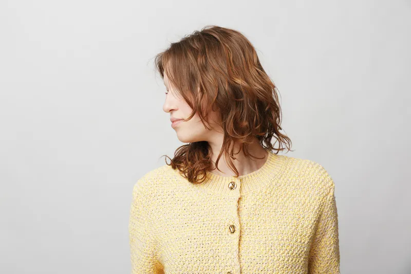 Young woman with curly brown hair wearing a yellow knitted sweater looks sideways on a light grey background. Casual style, natural beauty, and relaxed mood concept.