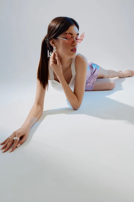 Young woman in stylish sunglasses and pastel outfit poses lying on white floor. Modern beauty and fashion concept with soft lighting and minimalistic background.