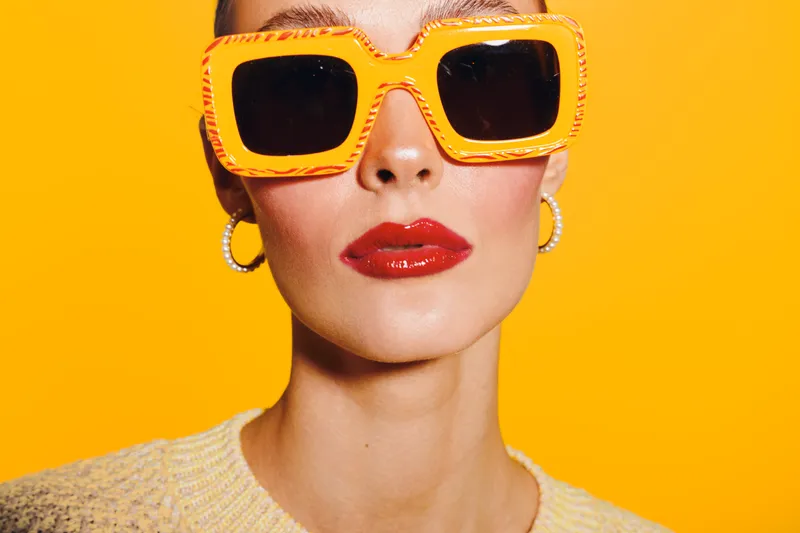 Stylish woman with bright orange sunglasses and red lipstick poses against vibrant yellow background. Fashion concept with bold colors and confident expression in studio lighting.