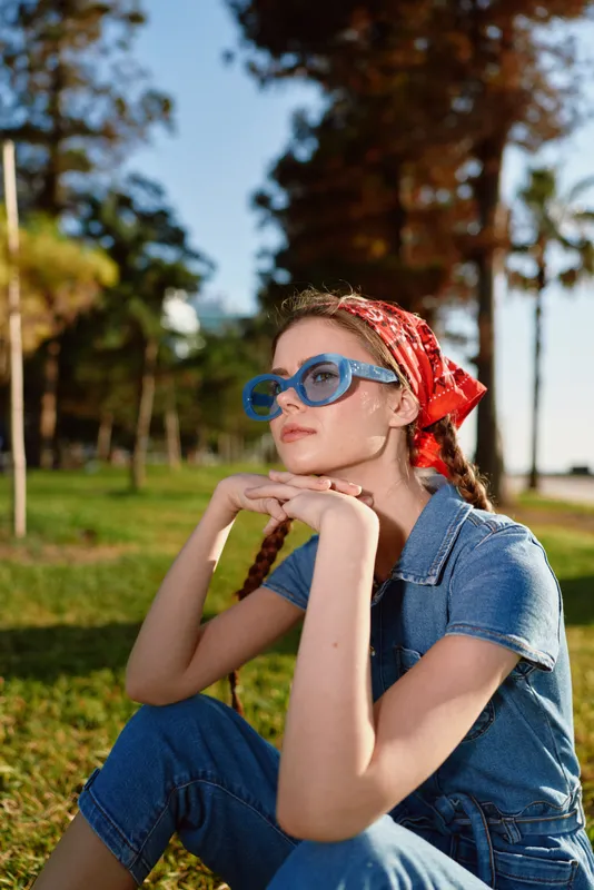lifestyle woman in modern boho-western denim street style wearing blue sunglasses and red bandana sitting outdoors in warm natural light with film color tones