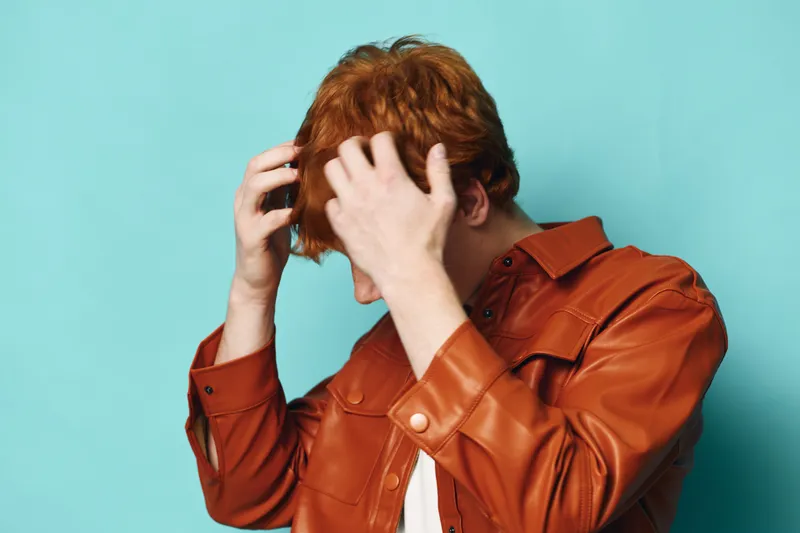 Young man with red hair in brown leather jacket adjusting hair on blue background. Casual style, fashion, hairstyle care and grooming for men with trendy look and confidence.