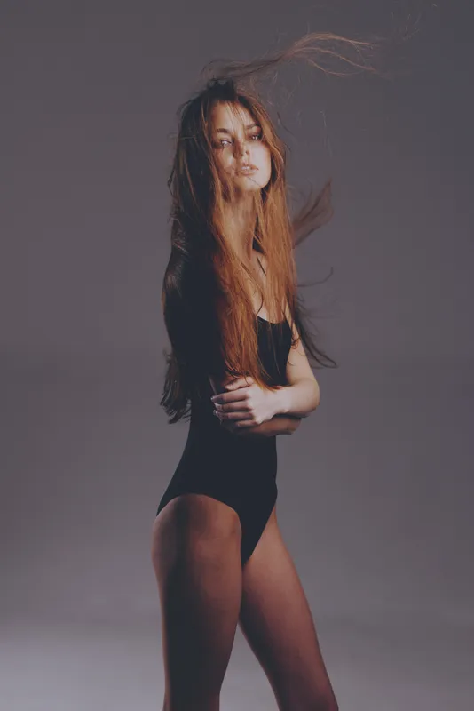 Young woman with flowing hair posing in black bodysuit against neutral background, dynamic and elegant style