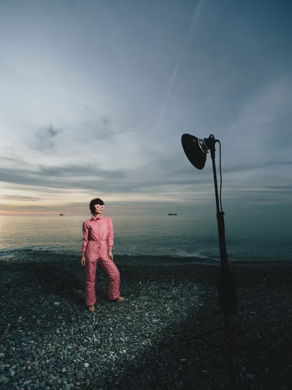 Portrait of a model in a pink jumpsuit standing on a pebbled beach beside a quiet sea with a large studio light on a stand creating dramatic outdoor fashion photography under an expansive evening sky