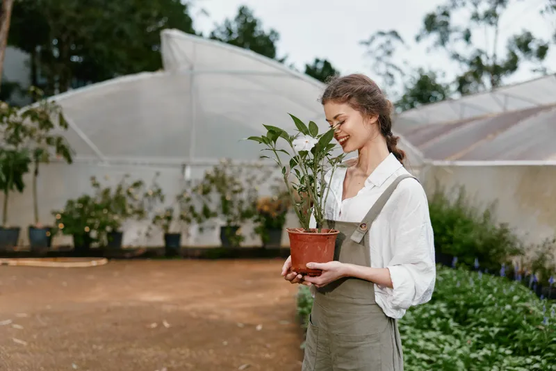 Young woman gardening in a greenhouse, holding a potted plant, dressed in casual overalls, exuding tranquility and joy in a verdant environment