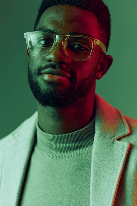 Stylish African man wearing glasses and a smart casual outfit, showcasing confidence in a green and pink lit background