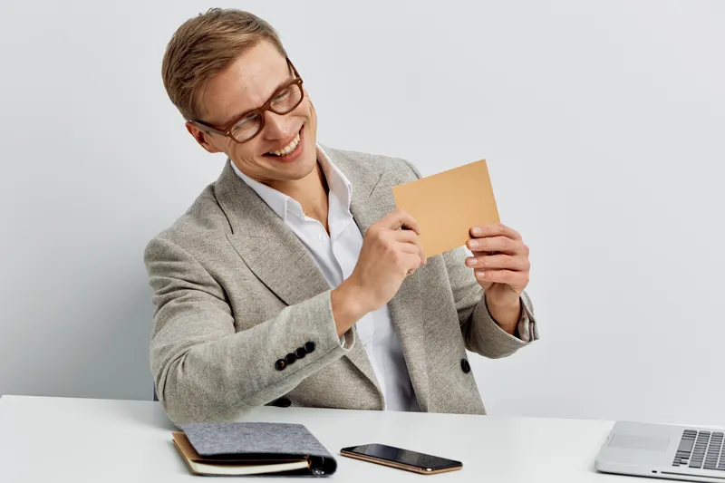 smiling man holding paper card wearing glasses gray jacket sitting at table with laptop mobile phone digital nomad work concept