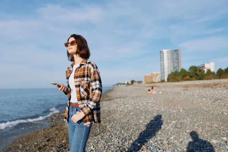 Woman on a pebbled beach with smartphone, wearing plaid shirt and denim jeans, sunglasses, coastal skyline in the distance under a clear sky, calm sea, natural light, casual outdoor portrait.