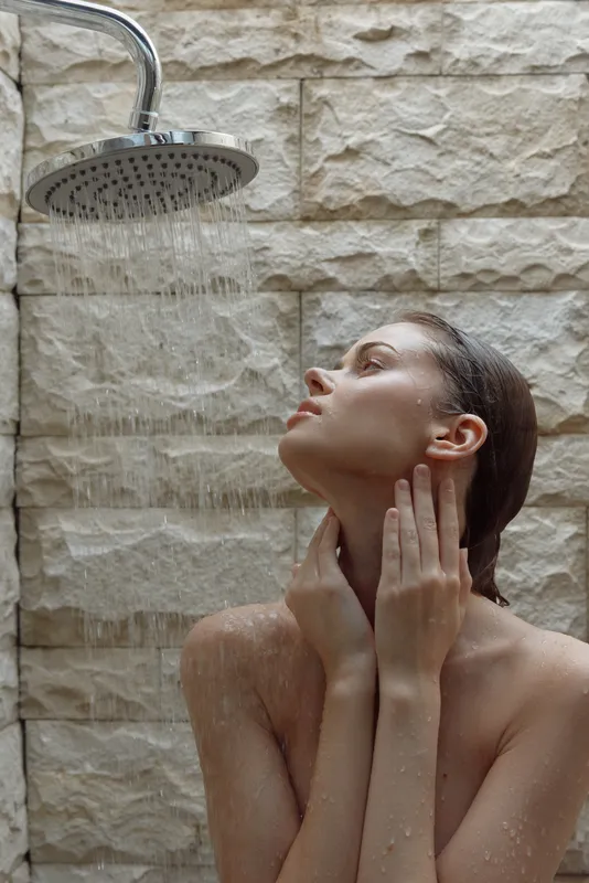 woman showering under rainfall showerhead with natural stone wall background, serene expression, wet hair, and minimalistic spa atmosphere, daily hygiene routine
