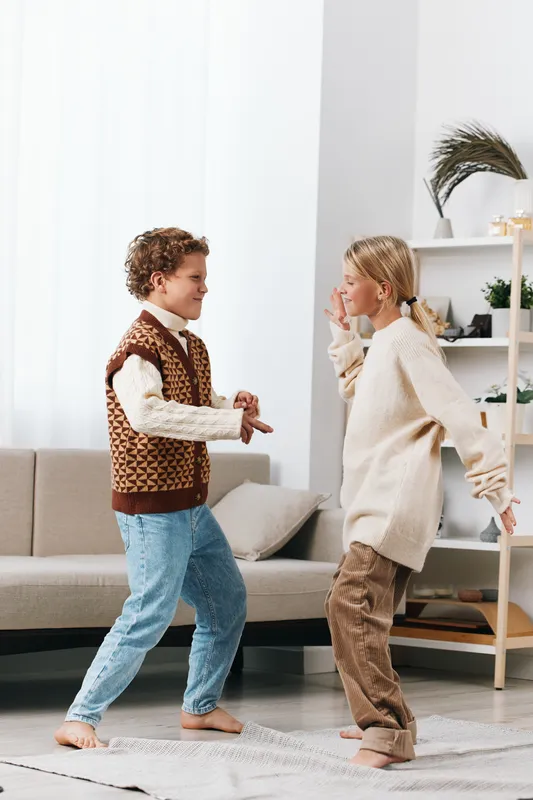 Two children dance together in a cozy living room, wearing warm outfits and barefoot on a soft rug, while soft natural light highlights their joyful movement.