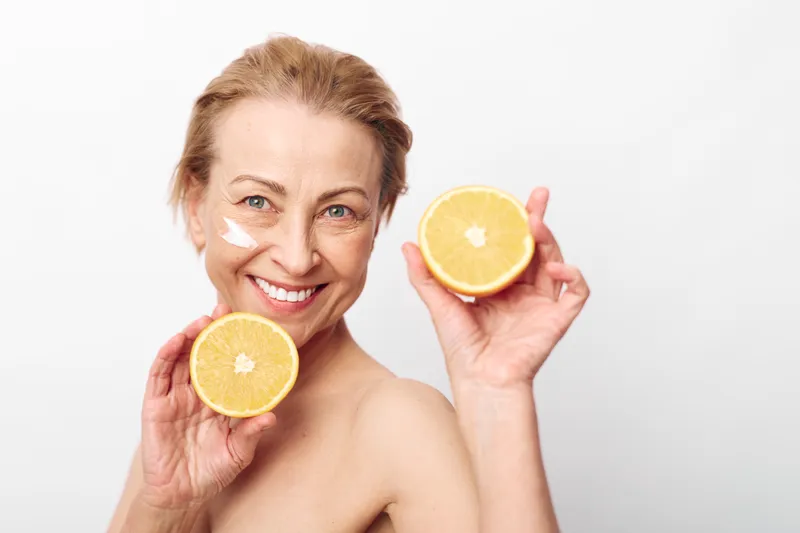 Smiling mature woman holds two halves of fresh grapefruit near face with cream on cheek. Concept of natural skincare, healthy lifestyle, citrus fruits, wellness, and beauty routine at home.