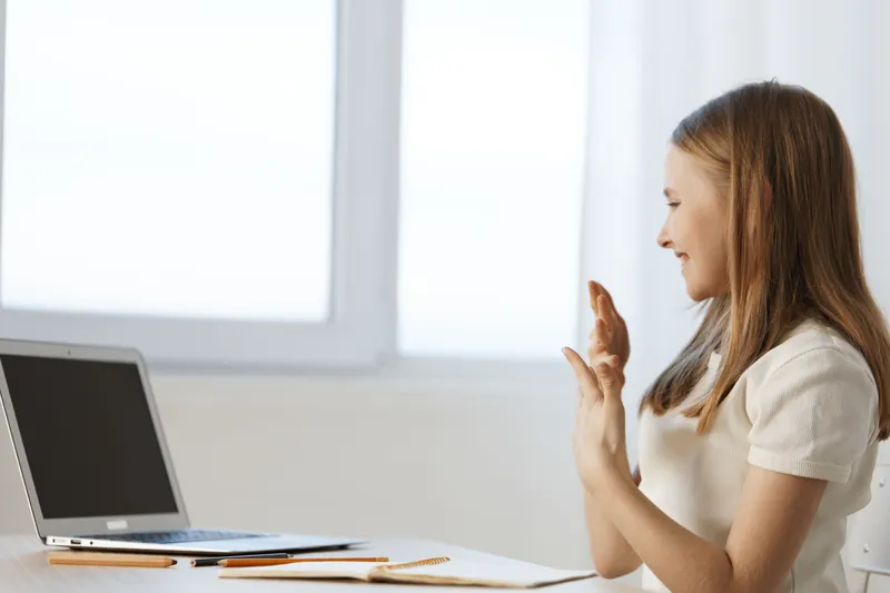 Cheerful young woman waving in front of a laptop, expressing joy during a video call, in a bright and minimalistic indoor setting, emphasizing connection and communication