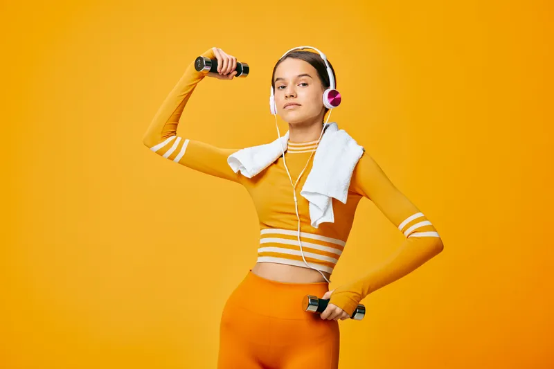 Young woman in orange activewear lifting small dumbbells with headphones and a towel over her shoulder, confident studio portrait on a vibrant orange monochrome background showcasing fitness and
