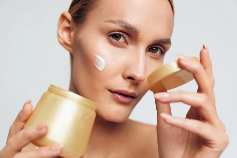 Woman applying white face cream from a golden jar, close up on a clean white background, skincare routine for beauty and wellness, soft studio lighting, self care