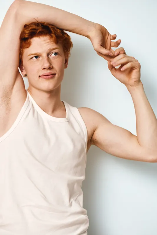 Young man with red hair stretching arms wearing sleeveless white shirt against plain light background. Casual posture with thoughtful expression for fitness or relaxation concept.