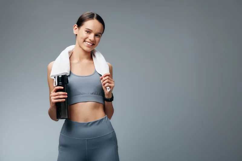 Fitness woman in a sports bra and leggings with a towel around her neck, holding a water bottle, standing in a studio ready for workout and training.