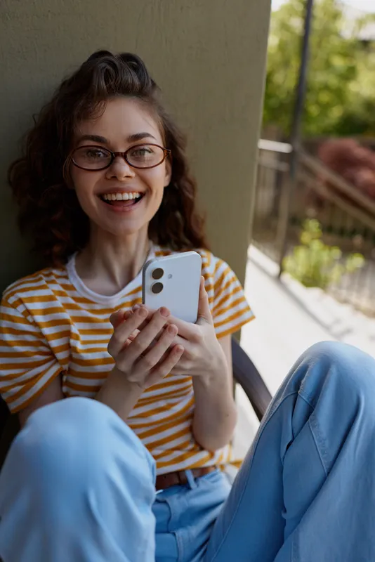 Cheerful woman at home, sitting on a balcony, smiling with phone in hand, striped tee, relaxed posture, sunlit moment, natural colors, contemporary lifestyle