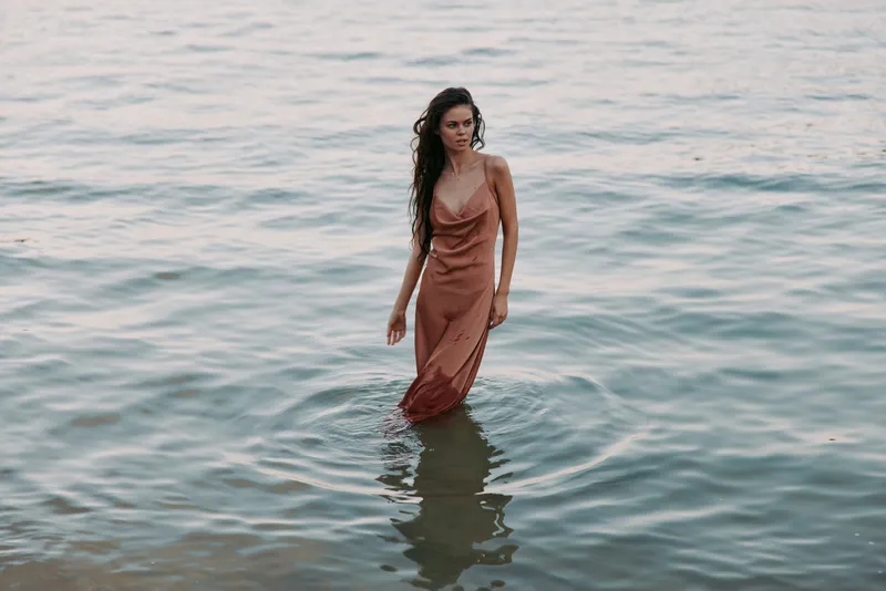 A lone woman stands in calm sea water wearing a soft rust dress, creating a reflective silhouette as gentle waves lap at her ankles under warm light.
