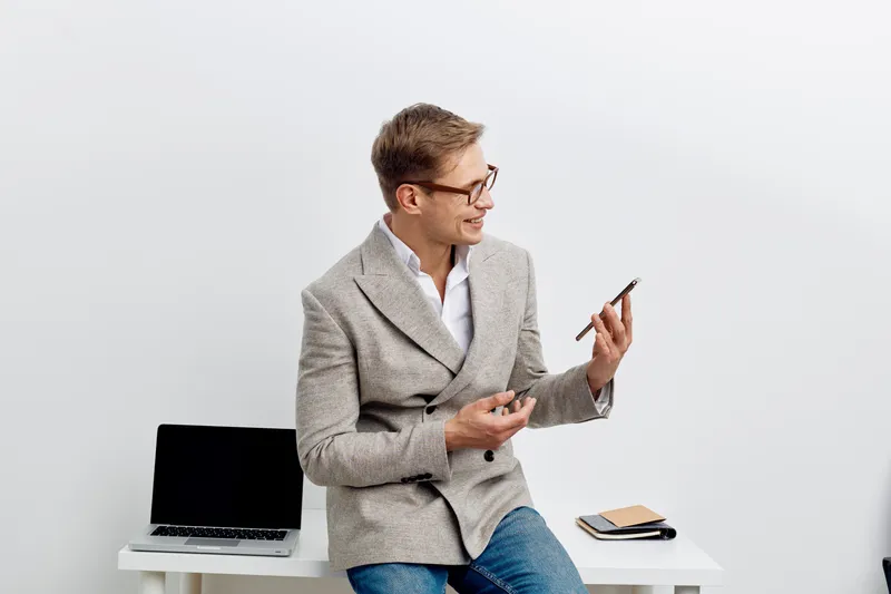 Professional smiling man in casual business attire sitting at a desk holding a smartphone, with open laptop and notebook nearby, indoors against a plain light background, modern workplace, lifestyle c